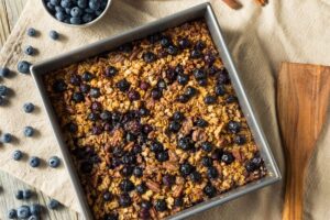 oatmeal bake postpartum freezer meal with blueberries on a table with a silver baking pan, a bowl of blueberries, and a wooden ladle